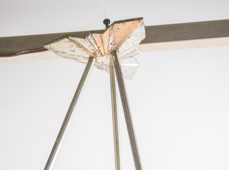 Pu&ograve; includere: Primo piano di un lampadario vintage in stile Art D&eacute;co. L'apparecchio presenta una parte superiore argentata a forma di stella con tre sottili aste argentate che si estendono verso il basso. Il design ricorda gli anni '20 e '30.
