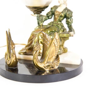 Puede incluir: Una l&aacute;mpara de mesa vintage con una figura de mujer verde y dorada sosteniendo un globo terr&aacute;queo. La base de la l&aacute;mpara est&aacute; hecha de m&aacute;rmol negro con una incrustaci&oacute;n de m&aacute;rmol blanco. Dos figuras de cisnes dorados est&aacute;n en la base.
