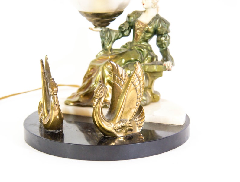 Puede incluir: Una l&aacute;mpara de mesa vintage con una figura de mujer verde y dorada sosteniendo un globo terr&aacute;queo. La base de la l&aacute;mpara est&aacute; hecha de m&aacute;rmol negro con una incrustaci&oacute;n de m&aacute;rmol blanco. Dos figuras de cisnes dorados est&aacute;n en la base.