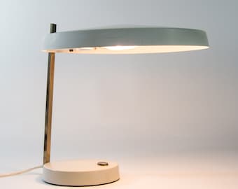 May include: A white desk lamp with a round, flat shade and a brass arm. The lamp has a white base and a small, round button on the side.