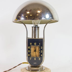 Peut inclure: Une lampe de table en chrome avec un abat-jour en forme de d&ocirc;me et un cadran d'horloge. Le cadran de l'horloge a des chiffres romains noirs et un fond blanc. La lampe est sur une base en marbre.