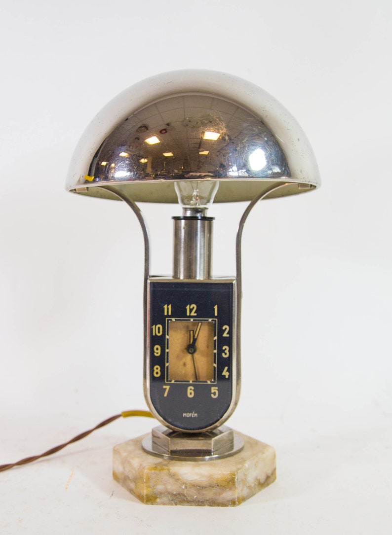 Peut inclure: Une lampe de table en chrome avec un abat-jour en forme de d&ocirc;me et un cadran d'horloge. Le cadran de l'horloge a des chiffres romains noirs et un fond blanc. La lampe est sur une base en marbre.