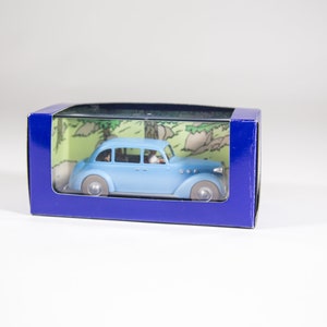 May include: A blue toy car in a clear plastic box with a blue background. The car is a vintage model with a white roof and chrome accents. The box has a cartoon image of a forest scene on the back.