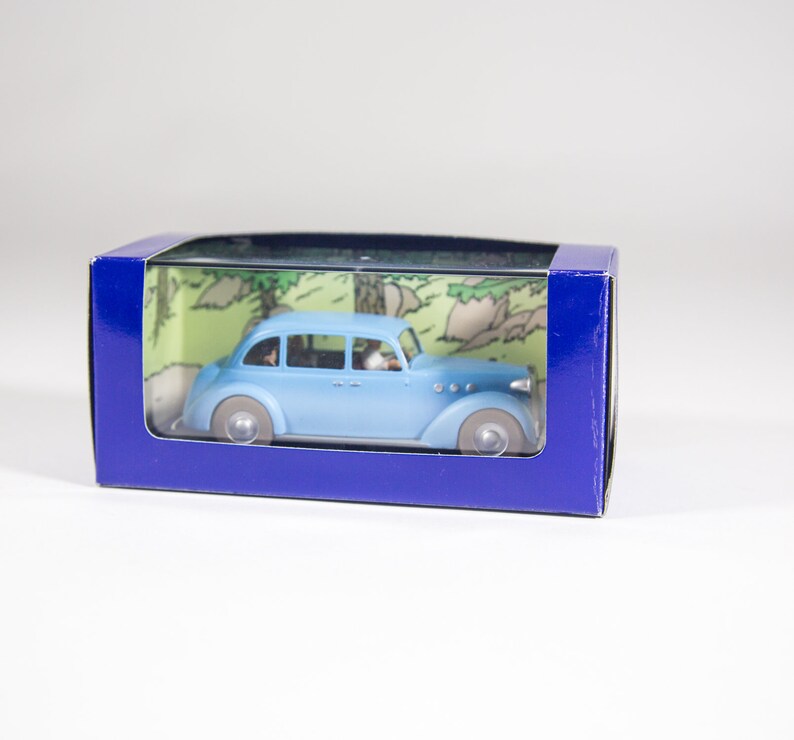 May include: A blue toy car in a clear plastic box with a blue background. The car is a vintage model with a white roof and chrome accents. The box has a cartoon image of a forest scene on the back.