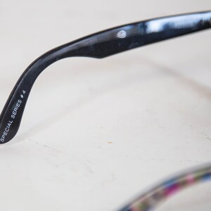 Puede incluir: Gafas de sol negras con acabado brillante en la patilla. El interior de la patilla tiene impreso el texto en blanco SPECIAL SERIES # 4. Un peque&ntilde;o logotipo circular es visible cerca de la bisagra. La montura incluye lentes de color verde oscuro.