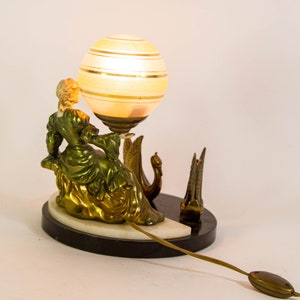 Puede incluir: Una l&aacute;mpara de mesa vintage con una figura de mujer verde y dorada sentada sobre un cisne. La l&aacute;mpara tiene una pantalla de globo blanco y dorado.