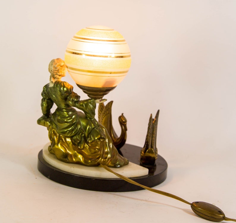 Puede incluir: Una l&aacute;mpara de mesa vintage con una figura de mujer verde y dorada sentada sobre un cisne. La l&aacute;mpara tiene una pantalla de globo blanco y dorado.