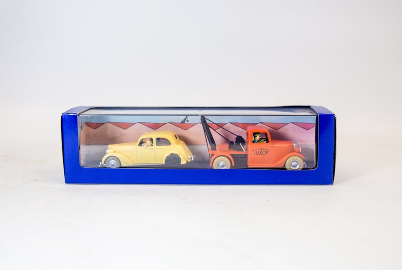 Puede incluir: Un coche amarillo siendo remolcado por una gr&uacute;a naranja. Ambos veh&iacute;culos est&aacute;n en una caja de pl&aacute;stico transparente con fondo azul. La gr&uacute;a tiene la palabra "LUXOR" impresa en el lateral.