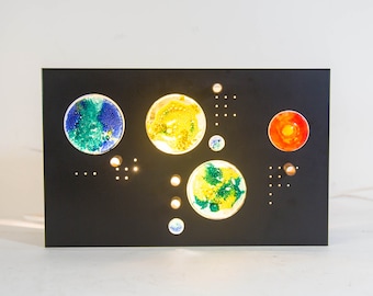 May include: A black rectangular wall art piece with cutouts of different coloured circles. The circles are filled with a mixture of green, blue, yellow, and orange. The piece is lit from behind, creating a glowing effect.