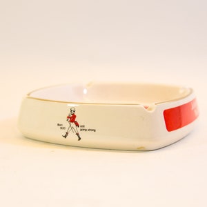 May include: White ceramic ashtray with a gold rim and a red accent. The ashtray features a graphic of a man in a red coat and the text "Born 1820 still going strong". The ashtray is square-shaped with rounded edges.
