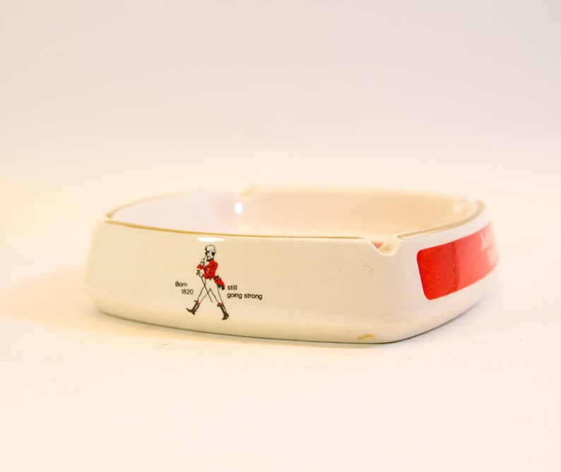 May include: White ceramic ashtray with a gold rim and a red accent. The ashtray features a graphic of a man in a red coat and the text "Born 1820 still going strong". The ashtray is square-shaped with rounded edges.