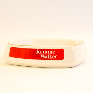 May include: White ceramic ashtray with a red rectangular label that reads "Johnnie Walker" in white text. The ashtray has a curved design with a small notch for resting cigarettes.