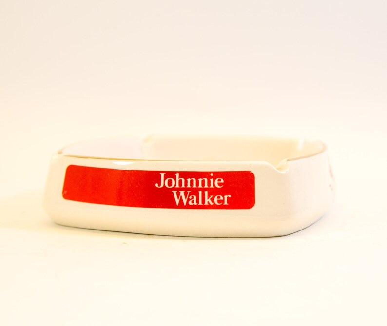 May include: White ceramic ashtray with a red rectangular label that reads "Johnnie Walker" in white text. The ashtray has a curved design with a small notch for resting cigarettes.