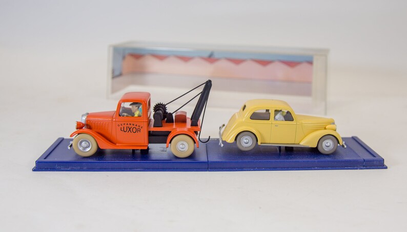 Puede incluir: Un cami&oacute;n de remolque rojo con un coche amarillo que est&aacute; siendo remolcado. El cami&oacute;n de remolque tiene la palabra "LUXOR" en el lateral. Ambos veh&iacute;culos est&aacute;n sobre una base de exposici&oacute;n azul.