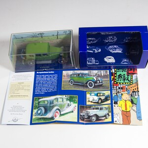 May include: A green and black model car in a clear plastic box with a blue and white box next to it. The blue box has a picture of a car on it. The boxes are on a white surface. The model car is a 1930s Dodge Brothers Coupe.