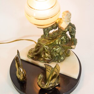 Puede incluir: Una l&aacute;mpara de mesa vintage con una figura esculpida dorada de una mujer sosteniendo una pantalla de l&aacute;mpara en forma de globo. La base es de m&aacute;rmol blanco y negro con dos figuras de cisnes doradas.