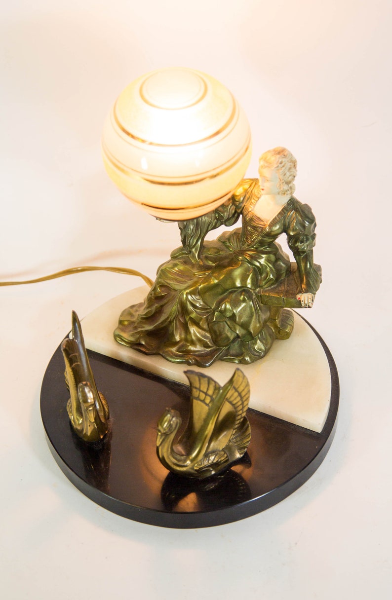 Puede incluir: Una l&aacute;mpara de mesa vintage con una figura esculpida dorada de una mujer sosteniendo una pantalla de l&aacute;mpara en forma de globo. La base es de m&aacute;rmol blanco y negro con dos figuras de cisnes doradas.