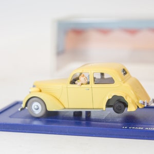 Puede incluir: Un coche de juguete amarillo siendo remolcado por un cami&oacute;n de juguete rojo con un brazo de gr&uacute;a negro. El coche tiene una peque&ntilde;a figura de una persona en el asiento del conductor. La base de la pantalla es azul con el texto "LE CRABE AUX PINCES D'OR" impreso en ella.