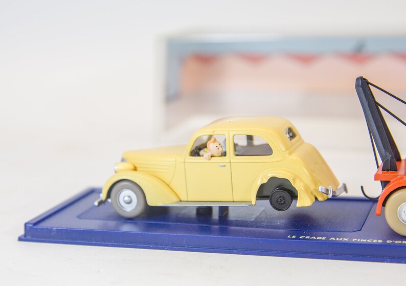 Puede incluir: Un coche de juguete amarillo siendo remolcado por un cami&oacute;n de juguete rojo con un brazo de gr&uacute;a negro. El coche tiene una peque&ntilde;a figura de una persona en el asiento del conductor. La base de la pantalla es azul con el texto "LE CRABE AUX PINCES D'OR" impreso en ella.