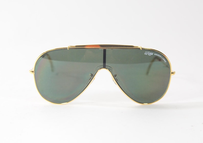 May include: Gold-framed aviator sunglasses with green lenses. The sunglasses have a 'Wings' logo on the right frame.