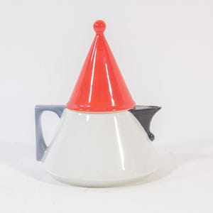 Op de afbeelding: Een witte keramische theepot met een rode conische deksel en een kleine rode knop. De handgreep is lichtblauw en de tuit is zwart. De theepot heeft een afgeronde body en een modern design.