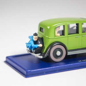 May include: A green toy car with a blue and white figure sitting on the back bumper. The car has a black base and the words "Le Lotus" are printed on the base.