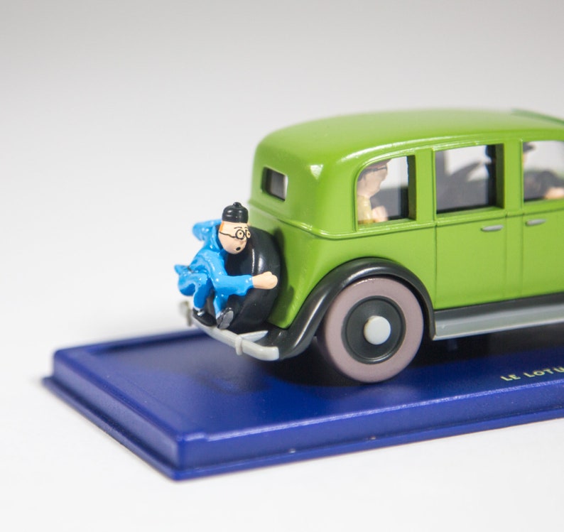 May include: A green toy car with a blue and white figure sitting on the back bumper. The car has a black base and the words "Le Lotus" are printed on the base.