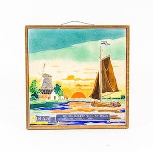 Könnte beinhalten: Eine antike holländische Fliese mit einer blauen und weißen Windmühle, einem Segelboot mit niederländischer Flagge und einem Sonnenaufgang über einem Gewässer. Die Fliese ist in Holz gerahmt und trägt den Text "1945 DE ORANJEZON SCHIJNT WEER OVER ONS VADERLAND."