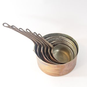 Op de afbeelding: Een set van zeven koperen saucepannen in verschillende maten, in elkaar genest. De saucepannen hebben lange handvatten en zijn gemaakt van glanzend, verweerd koper.
