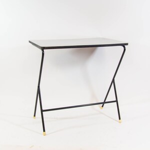 May include: A white table with a black metal frame and gold-coloured tips on the legs. The table has a simple, modern design.
