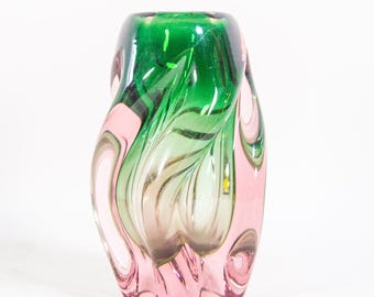 May include: A decorative glass vase with a unique, organic shape. The vase features a blend of colours, including pink, green, and clear glass. The top of the vase is a vibrant green, transitioning to pink and clear tones towards the base. The design is swirled and textured.