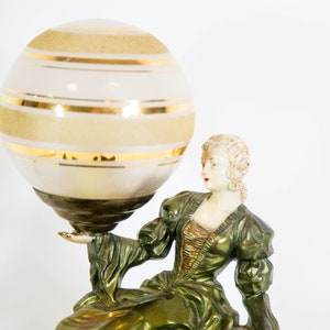 Puede incluir: Una l&aacute;mpara de mesa vintage con acabado verde y dorado. La l&aacute;mpara presenta una figura femenina sosteniendo un gran globo blanco con rayas doradas.