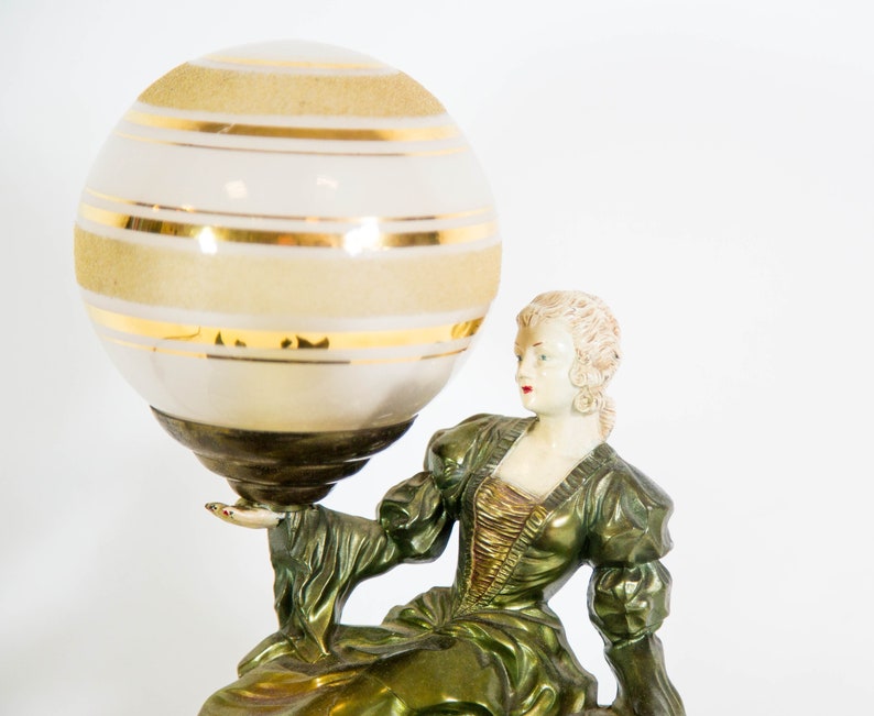 Puede incluir: Una l&aacute;mpara de mesa vintage con acabado verde y dorado. La l&aacute;mpara presenta una figura femenina sosteniendo un gran globo blanco con rayas doradas.