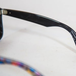 Puede incluir: Gafas de sol negras con lentes verde oscuro y acabado brillante. La patilla interior muestra el texto impreso en blanco WAYFARER Hand Made in Italy B CE RB2140 1049 50 22 3N. La otra patilla presenta un dise&ntilde;o colorido con m&uacute;ltiples patrones.