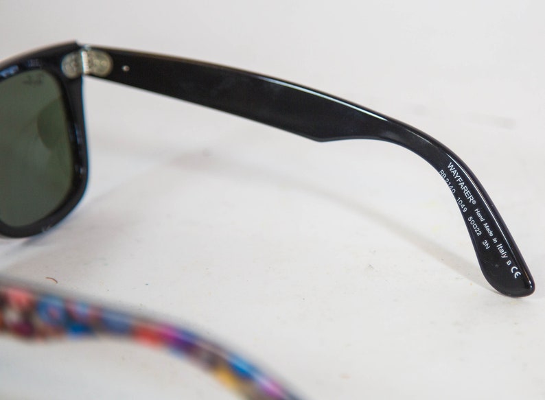 Puede incluir: Gafas de sol negras con lentes verde oscuro y acabado brillante. La patilla interior muestra el texto impreso en blanco WAYFARER Hand Made in Italy B CE RB2140 1049 50 22 3N. La otra patilla presenta un dise&ntilde;o colorido con m&uacute;ltiples patrones.