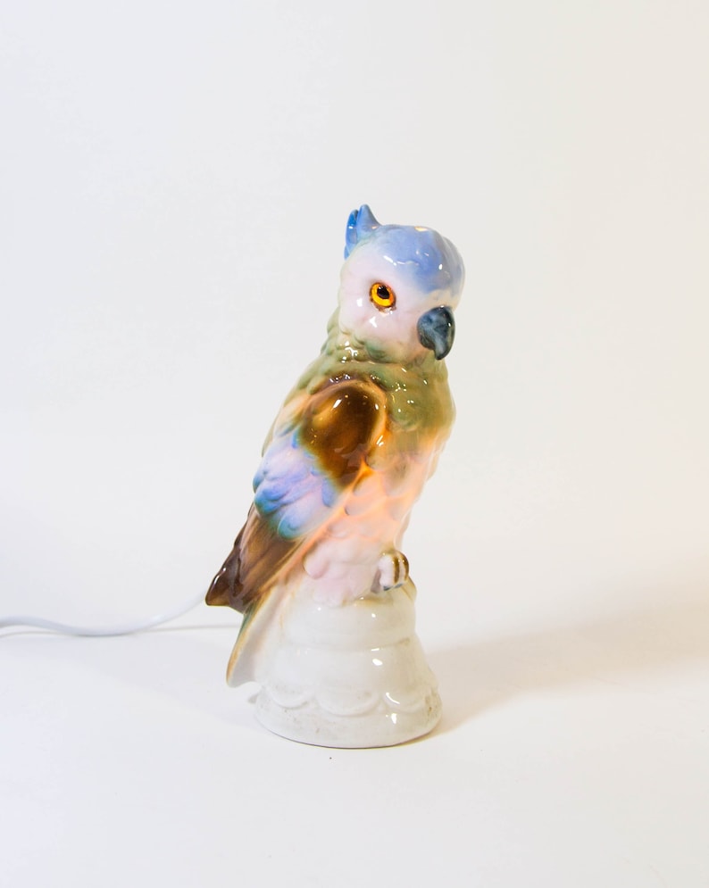 May include: A ceramic parrot nightlight with a blue, green, brown, and pink painted design. The parrot is perched on a white base with a cord.
