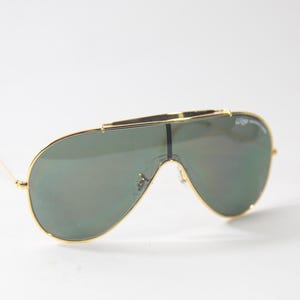 May include: Gold-framed aviator sunglasses with green mirrored lenses. The sunglasses have a thin metal frame and a bridge that connects the two lenses.