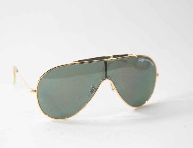 May include: Gold-framed aviator sunglasses with green mirrored lenses. The sunglasses have a thin metal frame and a bridge that connects the two lenses.