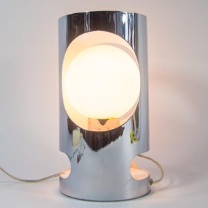 Puede incluir: Lámpara de mesa cilíndrica cromada con un gran recorte circular que revela una fuente de luz esférica brillante. La lámpara tiene un diseño moderno y una superficie reflectante. Un cable de alimentación sale de la base. Mide unos 25 cm de alto.