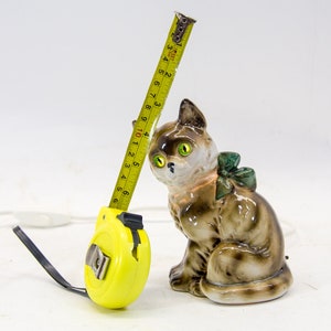 May include: A yellow tape measure with a brown and white ceramic cat figurine. The cat is wearing a green bow and has green eyes. The tape measure is extended and reads 456 34 4 5 6 7 8 9 10 1 2 3 4 5 6.