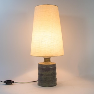 Op de afbeelding: Een vintage tafellamp met een getextureerde, cilindrische voet in groen- en bruintinten. De lamp heeft een beige, conische lampenkap en een zwart netsnoer met een aan/uit-schakelaar. De lamp is een decoratieve verlichtingsarmatuur.