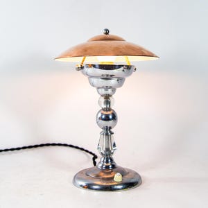 May include: A vintage desk lamp with a copper shade and a chrome base. The lamp features a decorative glass and chrome stem, and a round base. The lamp is turned on, emitting a warm glow. The lamp's cord is black.