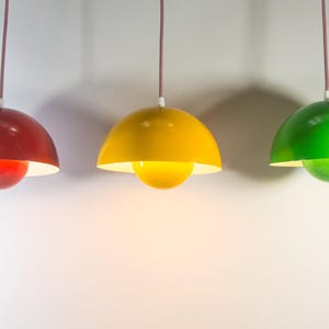 Op de afbeelding: Drie hanglampen in rood, geel en groen. Elke lamp heeft een afgeronde, koepelvormige kap en een bolvormig bodemelement. De lampen hangen aan dunne, bruine snoeren tegen een witte achtergrond. De lampen zijn gelijkmatig verdeeld.