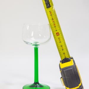 May include: A yellow tape measure with a black and yellow handle is next to a green stemmed wine glass. The tape measure is extended to 40 cm.