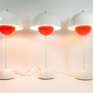 Op de afbeelding: Drie tafellampen met een witte koepelvormige kap en een kleiner oranje halfrond element eronder. Elke lamp heeft een slanke witte steel en een witte halfronde voet, met een wit netsnoer dat uit de onderkant van elke eenheid komt.