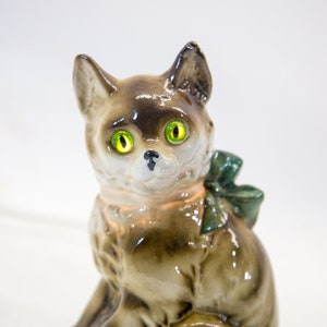 May include: A ceramic figurine of a cat with green eyes and a green bow. The cat is sitting with its head tilted to the side.