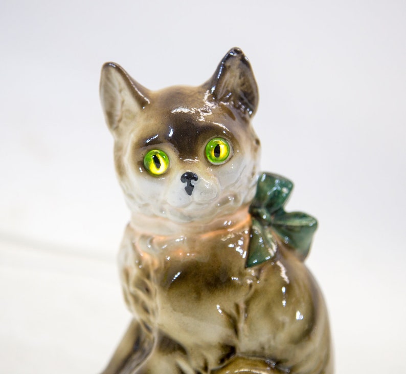 May include: A ceramic figurine of a cat with green eyes and a green bow. The cat is sitting with its head tilted to the side.