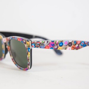 Puede incluir: Gafas de sol Ray-Ban con una montura que presenta un patr&oacute;n multicolor de peque&ntilde;os iconos circulares. Las lentes son de color verde oscuro y el interior de la montura y las patillas son de color negro s&oacute;lido. El logo Ray-Ban est&aacute; impreso en blanco.