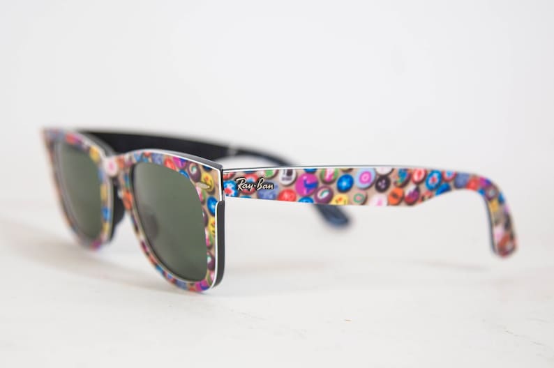 Puede incluir: Gafas de sol Ray-Ban con una montura que presenta un patr&oacute;n multicolor de peque&ntilde;os iconos circulares. Las lentes son de color verde oscuro y el interior de la montura y las patillas son de color negro s&oacute;lido. El logo Ray-Ban est&aacute; impreso en blanco.