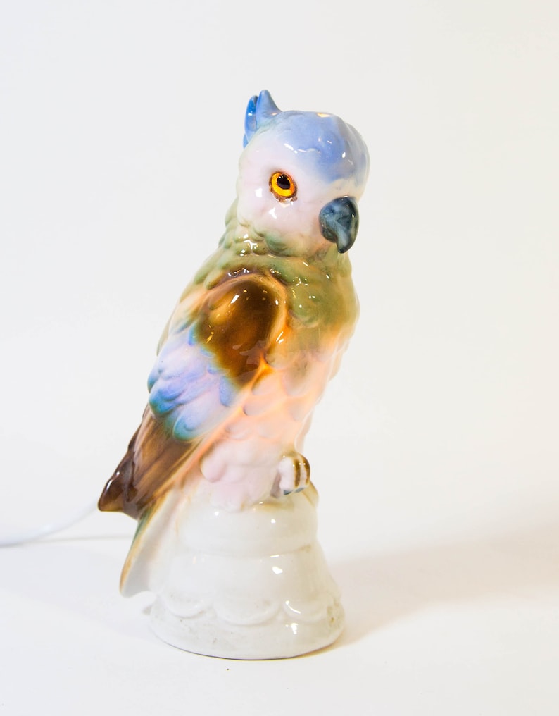 May include: A ceramic parrot figurine with a blue, green, brown, and pink paint scheme. The parrot is perched on a white base.
